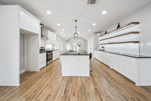 kitchen with a kitchen island, stainless steel gas stove, white cabinetry, hanging light fixtures, and wall chimney exhaust hood