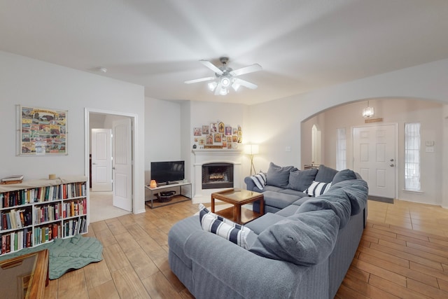 living area with arched walkways, a fireplace, light wood-style flooring, and ceiling fan