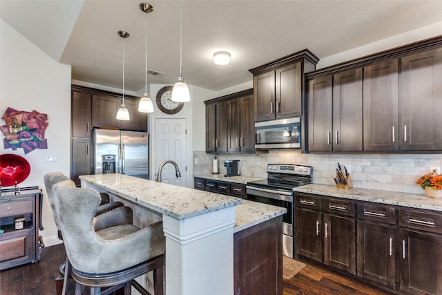 kitchen with an island with sink, hanging light fixtures, light stone counters, dark brown cabinetry, and stainless steel appliances