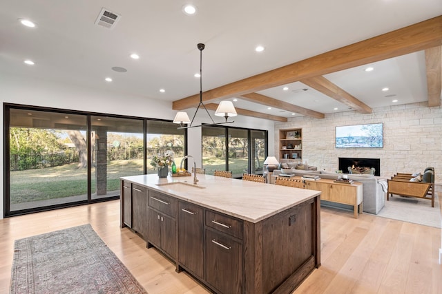 kitchen featuring pendant lighting, an island with sink, sink, dark brown cabinetry, and light stone counters