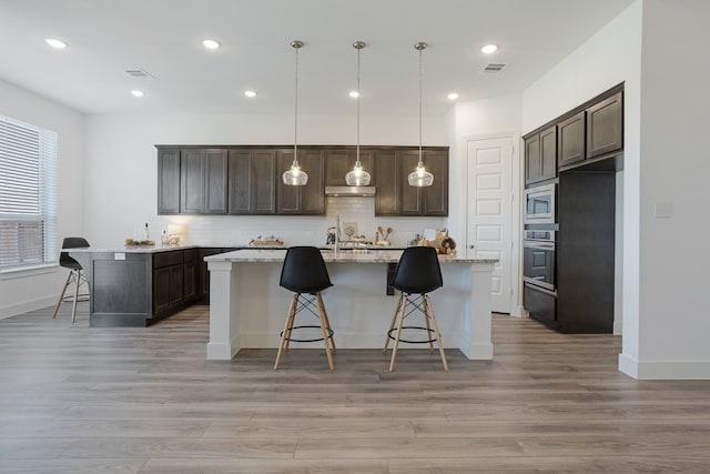 kitchen with light stone counters, dark brown cabinets, hanging light fixtures, a center island with sink, and stainless steel appliances