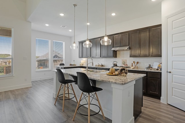 kitchen featuring pendant lighting, dark brown cabinetry, light stone counters, and a center island with sink