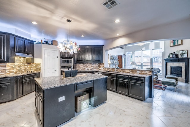 kitchen with tasteful backsplash, an island with sink, hanging light fixtures, light stone counters, and stainless steel appliances