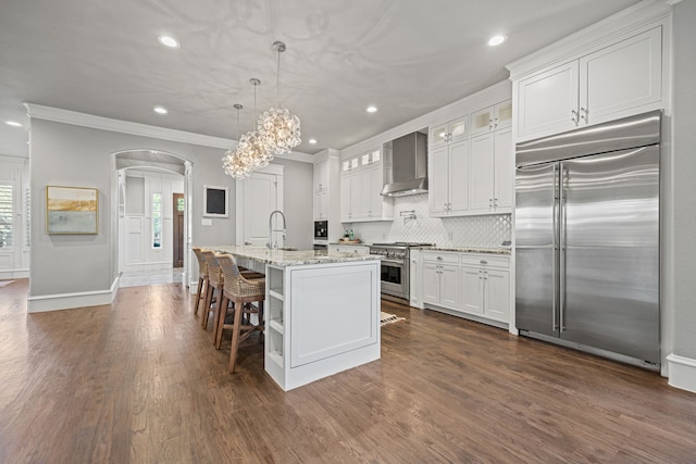 kitchen with pendant lighting, white cabinetry, premium appliances, a center island with sink, and wall chimney exhaust hood