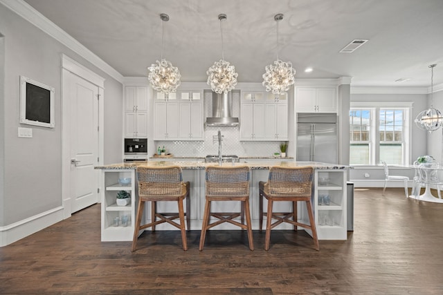 kitchen featuring built in fridge, pendant lighting, a chandelier, a kitchen island with sink, and wall chimney range hood