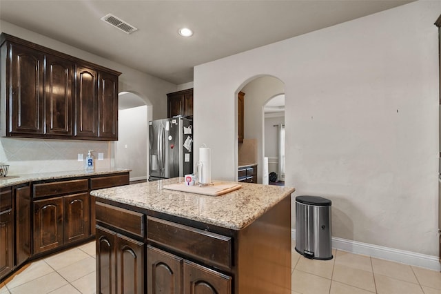 kitchen with stainless steel refrigerator with ice dispenser, dark brown cabinetry, tasteful backsplash, light tile patterned floors, and a kitchen island