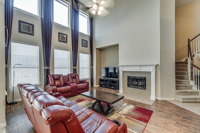 living room featuring a ceiling fan, a tile fireplace, stairway, wood finished floors, and a high ceiling