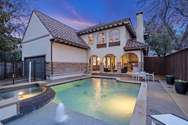 back of property at dusk featuring a patio, a chimney, a pool with connected hot tub, stone siding, and a fenced backyard