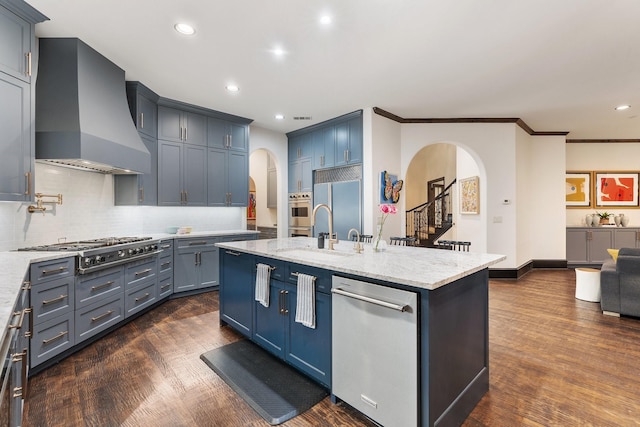 kitchen with arched walkways, appliances with stainless steel finishes, a kitchen island with sink, a sink, and wall chimney exhaust hood