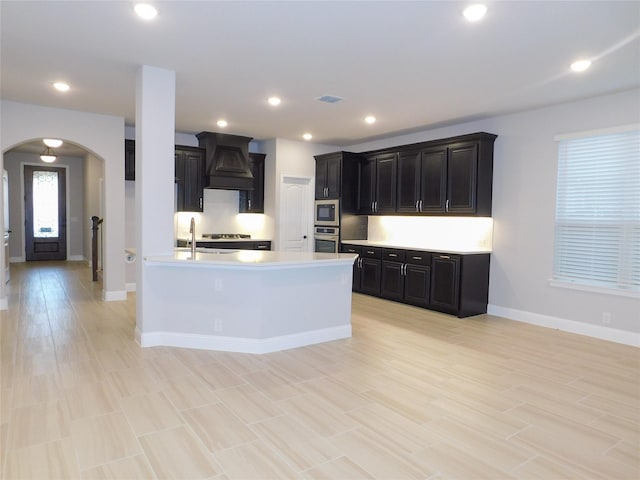 kitchen featuring black microwave, stainless steel oven, custom range hood, a center island with sink, and light hardwood / wood-style flooring