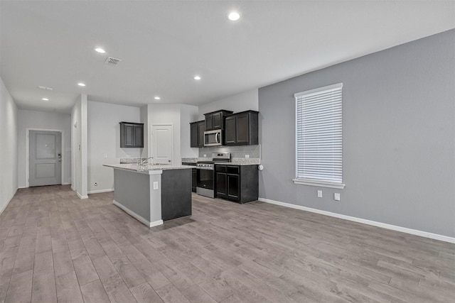 kitchen featuring light hardwood / wood-style flooring, stainless steel appliances, light stone countertops, a kitchen island with sink, and decorative backsplash