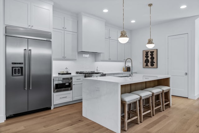 kitchen featuring wall chimney exhaust hood, stainless steel built in refrigerator, a center island with sink, and white cabinets