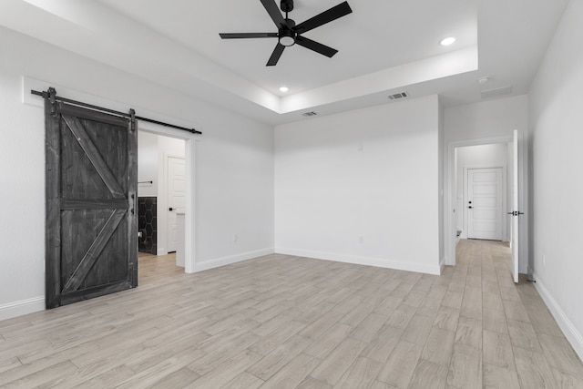 empty room with a tray ceiling, a barn door, ceiling fan, and light wood-type flooring