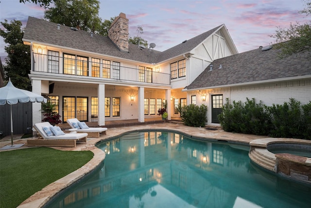 back house at dusk featuring a balcony, a pool with hot tub, and a patio