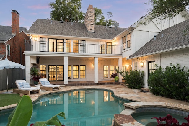 back house at dusk featuring a swimming pool with hot tub, a balcony, and a patio area