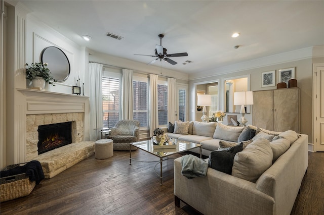 living room with crown molding, a stone fireplace, dark wood-type flooring, and ceiling fan