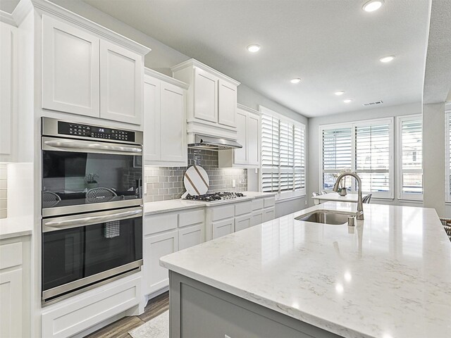 kitchen with sink, white cabinetry, light stone counters, stainless steel appliances, and wall chimney range hood