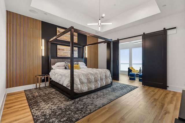 bedroom featuring ceiling fan, a tray ceiling, a barn door, and light hardwood / wood-style flooring