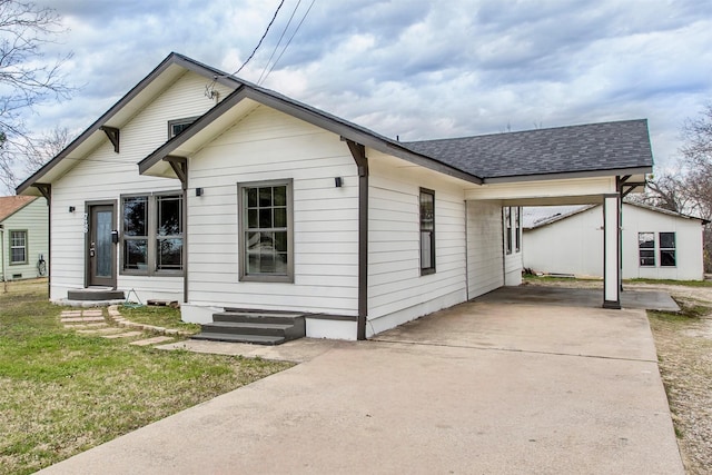 bungalow-style home with a shingled roof, concrete driveway, entry steps, an attached carport, and a front lawn