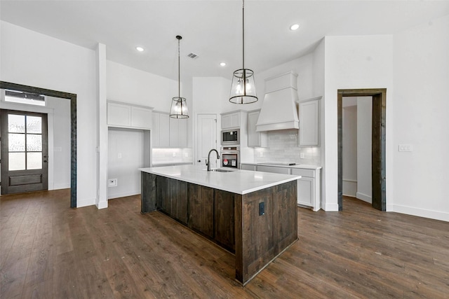 kitchen with appliances with stainless steel finishes, decorative light fixtures, tasteful backsplash, custom exhaust hood, and a kitchen island with sink