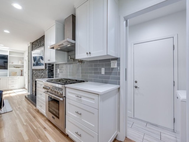 kitchen with wall chimney range hood, stainless steel stove, white cabinetry, tasteful backsplash, and light wood-type flooring