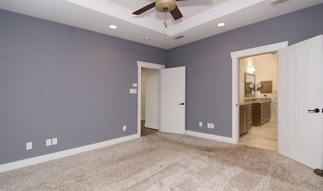 unfurnished bedroom with a tray ceiling, ensuite bath, light colored carpet, and ceiling fan