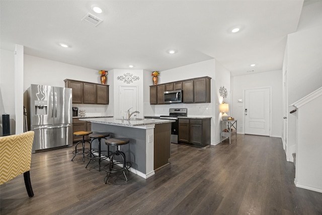 kitchen with appliances with stainless steel finishes, a kitchen island with sink, dark brown cabinetry, light stone countertops, and dark hardwood / wood-style flooring