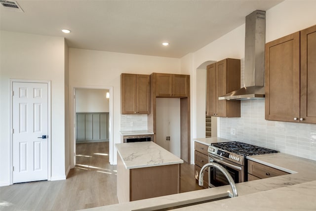 kitchen featuring wall chimney range hood, tasteful backsplash, light stone countertops, stainless steel range with gas cooktop, and light wood-type flooring