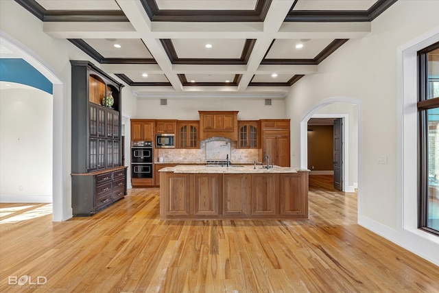 kitchen with built in microwave, tasteful backsplash, an island with sink, double oven, and beamed ceiling