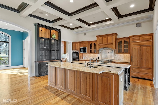 kitchen with appliances with stainless steel finishes, coffered ceiling, light stone countertops, a center island with sink, and light wood-type flooring