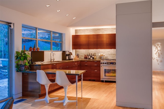 kitchen featuring sink, stainless steel stove, light hardwood / wood-style flooring, backsplash, and vaulted ceiling