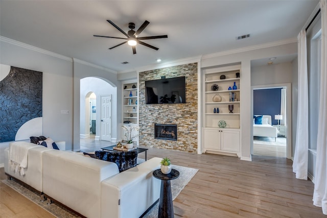 living room with crown molding, a stone fireplace, built in features, and light hardwood / wood-style flooring