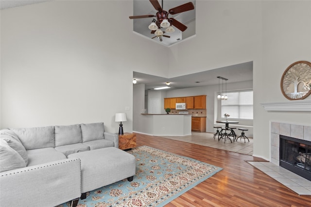 living room featuring a high ceiling, a tile fireplace, ceiling fan with notable chandelier, and light hardwood / wood-style floors