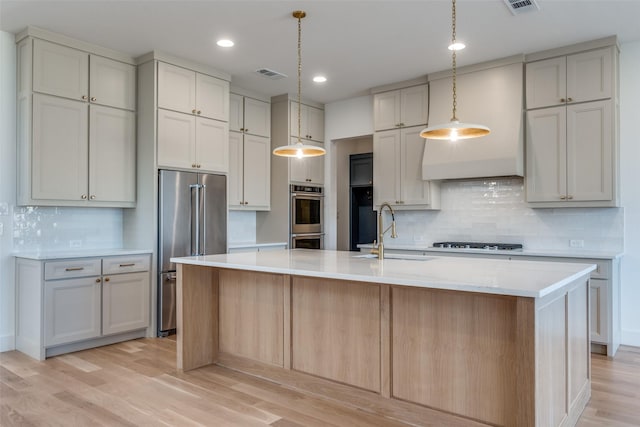 kitchen with sink, decorative light fixtures, a kitchen island with sink, stainless steel appliances, and light hardwood / wood-style floors