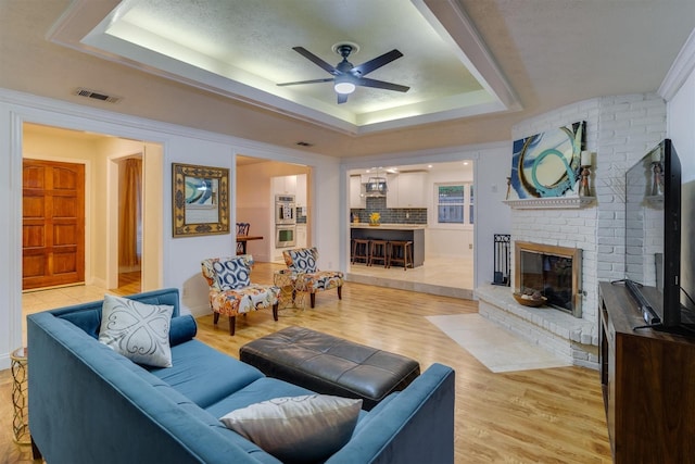 living room featuring ceiling fan, a fireplace, a tray ceiling, and light wood-type flooring