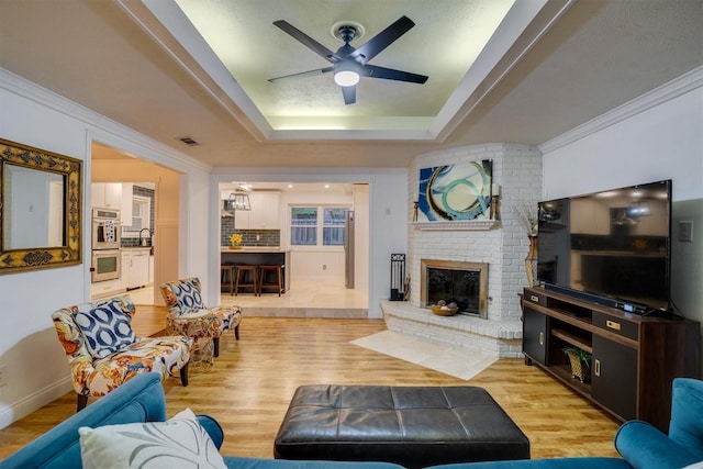 living room featuring light hardwood / wood-style flooring, ceiling fan, a tray ceiling, a fireplace, and ornamental molding