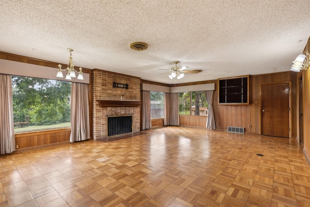 unfurnished living room featuring ceiling fan with notable chandelier, wood walls, light parquet floors, a brick fireplace, and a textured ceiling