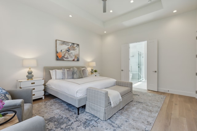 bedroom with ceiling fan, a tray ceiling, light wood-type flooring, and ensuite bath