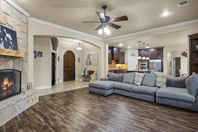 living room featuring a stone fireplace, sink, ceiling fan, crown molding, and dark wood-type flooring