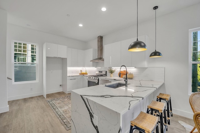 kitchen with wall chimney range hood, hanging light fixtures, gas stove, white cabinets, and kitchen peninsula