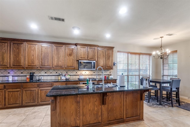 kitchen with sink, light tile patterned floors, dark stone countertops, a center island with sink, and decorative backsplash