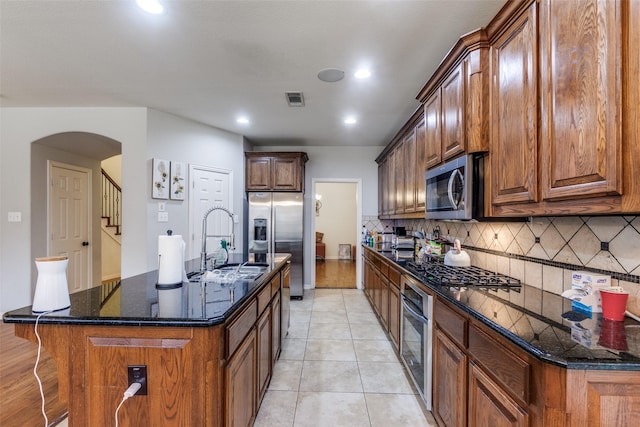 kitchen with sink, tasteful backsplash, dark stone counters, an island with sink, and stainless steel appliances