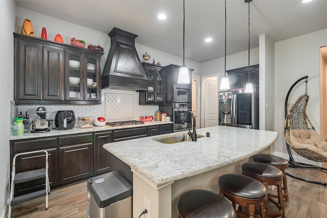 kitchen featuring sink, appliances with stainless steel finishes, dark brown cabinets, a center island with sink, and custom exhaust hood