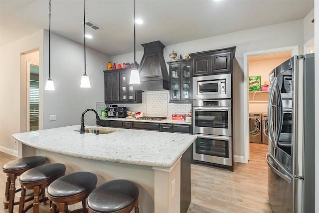 kitchen with sink, washer and clothes dryer, a kitchen island with sink, stainless steel appliances, and custom exhaust hood