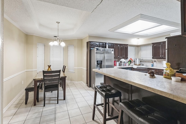 kitchen with light tile patterned floors, stainless steel fridge, dark brown cabinets, a tray ceiling, and decorative light fixtures