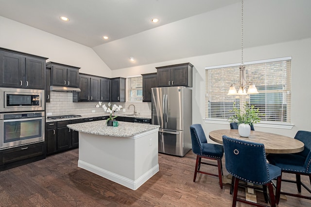 kitchen with appliances with stainless steel finishes, dark hardwood / wood-style floors, a center island, light stone countertops, and vaulted ceiling