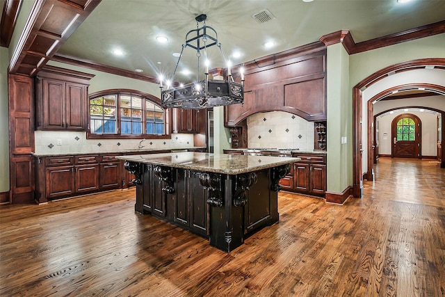 kitchen with dark wood-type flooring, dark brown cabinets, a kitchen island, pendant lighting, and light stone countertops