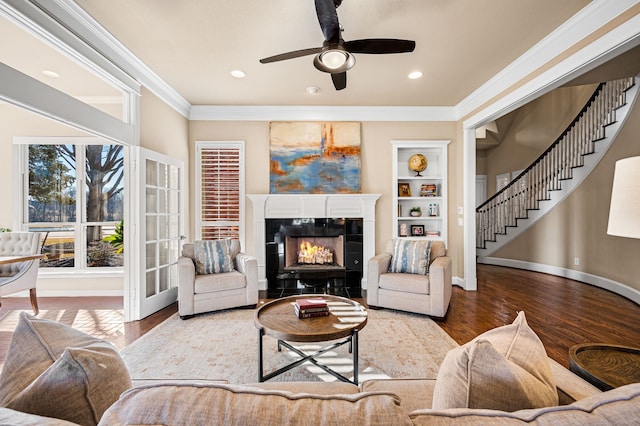 living room with a tile fireplace, built in features, wood-type flooring, ornamental molding, and ceiling fan