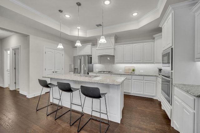 kitchen with appliances with stainless steel finishes, white cabinetry, light stone counters, a tray ceiling, and a center island with sink