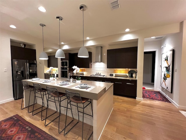 kitchen with appliances with stainless steel finishes, a kitchen island with sink, hanging light fixtures, wall chimney exhaust hood, and light wood-type flooring